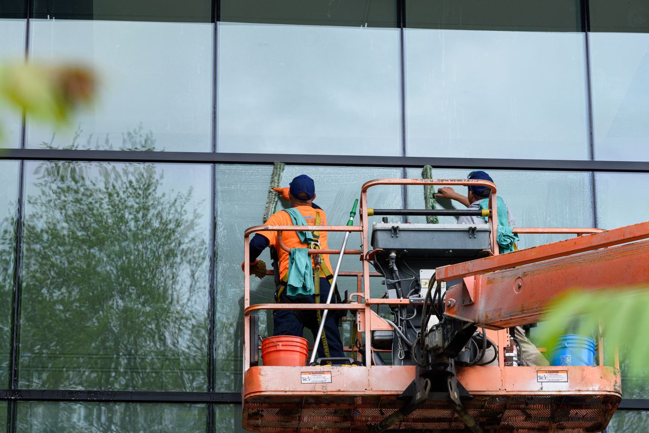Two workers washing windows on a glass building facade with a lift platform, improving the building's appearance.